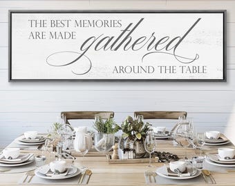May include: A rectangular wall sign with a dark frame and a white background featuring the text "THE BEST MEMORIES ARE MADE gathered AROUND THE TABLE" in a mix of block and cursive grey lettering, displayed above a wooden dining table.