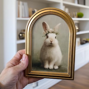 May include: A framed print featuring a white rabbit wearing a floral crown. The artwork is set within a gold-colored, arched frame. The rabbit is the focal point, set against a soft, muted background.