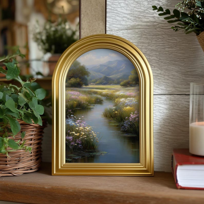 Pode incluir: Uma pintura de paisagem emoldurada com uma moldura dourada arqueada. A obra de arte retrata uma serena cena de rio com flores silvestres coloridas, &aacute;rvores e montanhas ao fundo. A pintura &eacute; exibida em uma superf&iacute;cie de madeira.