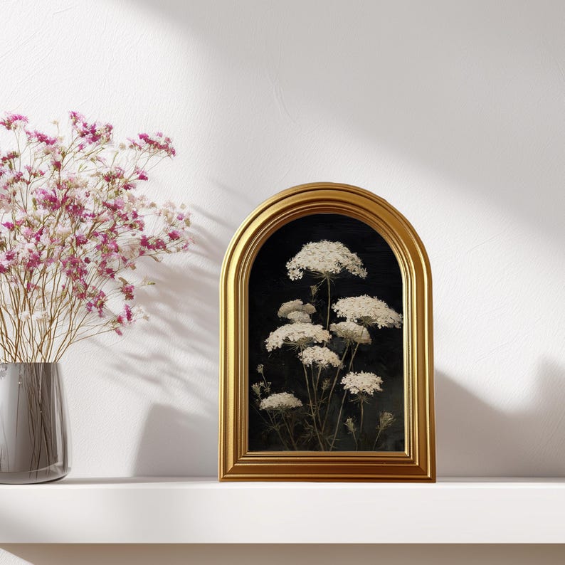 May include: A framed botanical print featuring white Queen Anne's Lace flowers against a dark background. The gold-colored frame has an arched top. A vase of pink flowers sits to the left of the framed print.