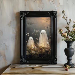 May include: A framed art print featuring two ghost figures in a field of flowers. The ghosts are draped in white sheets with black eye holes. The black frame has ornate detailing. The artwork is displayed on a wooden surface.