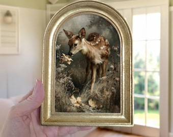 May include: A small, arched gold-toned frame containing a detailed print of a young spotted deer standing among dried grasses and fallen autumn leaves. The deer is depicted in profile, looking downward toward the foliage in a muted, natural setting.