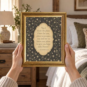 May include: A rectangular gold-toned frame containing a dark gray background with a white floral vine pattern. Centered is a cream-colored scalloped label featuring the text: For I know the plans I have for you, plans to prosper you and not to harm you, plans to give you hope and a future. Jeremiah 29:11.