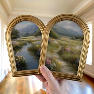 May include: Two framed landscape paintings with gold arched frames. The paintings depict a serene meadow scene with a river and mountains in the background. The artwork features a variety of colors, including green, blue, and yellow.