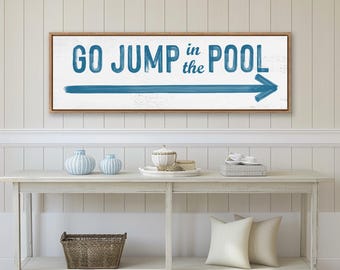May include: A rectangular wall sign with a white background and a wooden frame. The text reads "GO JUMP in the POOL" in blue, brush-stroke style lettering. Below the text is a long, horizontal blue arrow pointing toward the right side.