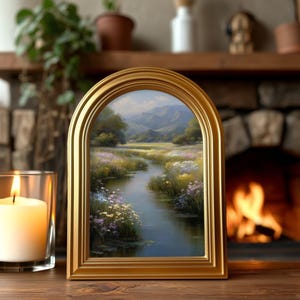 Pode incluir: Uma pintura de paisagem emoldurada com uma moldura dourada arqueada. A obra retrata uma serena cena de rio com montanhas ao fundo, vegeta&ccedil;&atilde;o exuberante e flores silvestres coloridas. Uma vela acesa em um casti&ccedil;al de vidro est&aacute; &agrave; esquerda.
