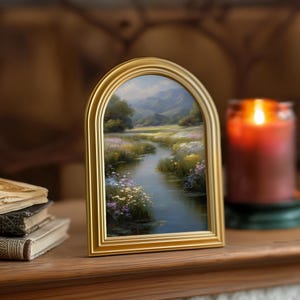 Pode incluir: Uma moldura dourada em forma de arco com uma pintura de paisagem de um rio que flui por um campo de flores silvestres. A pintura tem uma qualidade serena, com montanhas ao fundo. Uma vela acesa &eacute; vis&iacute;vel.