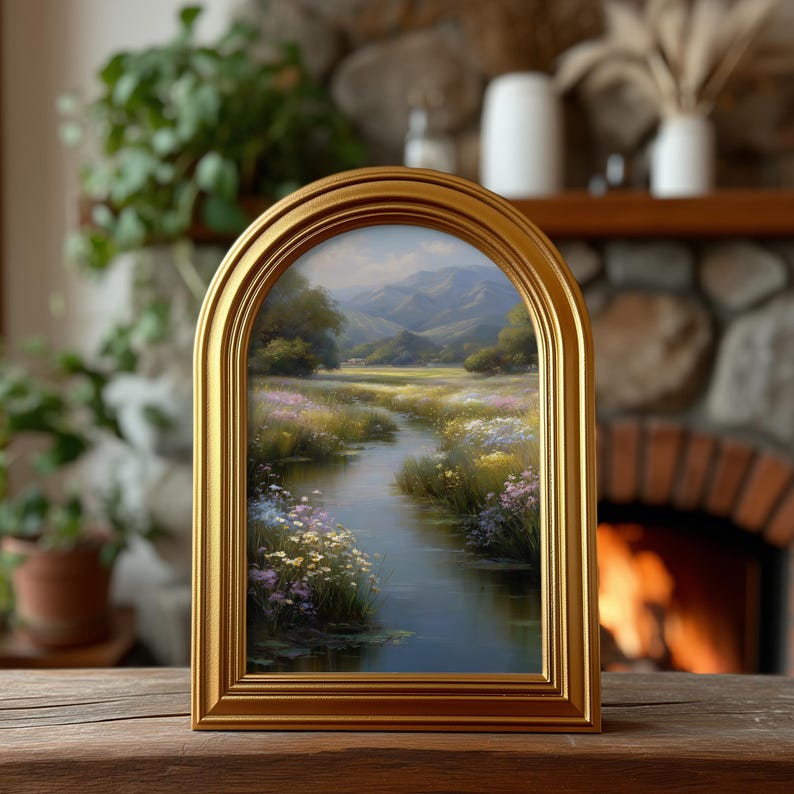 Pode incluir: Uma pintura de paisagem emoldurada em ouro com uma moldura arqueada. A obra de arte retrata uma cena de rio serena com montanhas, &aacute;rvores e flores silvestres. A moldura dourada &eacute; exibida em uma superf&iacute;cie de madeira.