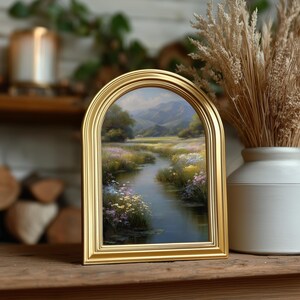 Pode incluir: Uma pintura de paisagem emoldurada com uma moldura dourada arqueada. A obra de arte retrata uma serena cena de rio com flores silvestres coloridas, &aacute;rvores e montanhas ao fundo. A pintura &eacute; exibida em uma superf&iacute;cie de madeira, ao lado de um vaso com plantas secas.