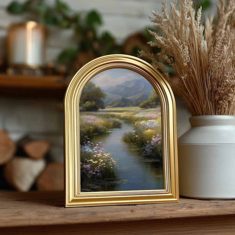 Pode incluir: Uma pintura de paisagem emoldurada com uma moldura dourada arqueada. A obra de arte retrata uma serena cena de rio com flores silvestres coloridas, &aacute;rvores e montanhas ao fundo. A pintura &eacute; exibida em uma superf&iacute;cie de madeira, ao lado de um vaso com plantas secas.