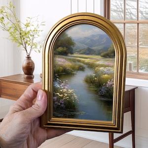 Pode incluir: Uma pintura de paisagem emoldurada com uma moldura dourada arqueada. A obra retrata uma serena cena de rio com flores silvestres coloridas, vegeta&ccedil;&atilde;o exuberante e montanhas distantes sob um c&eacute;u brilhante. A moldura adiciona um toque de eleg&acirc;ncia.