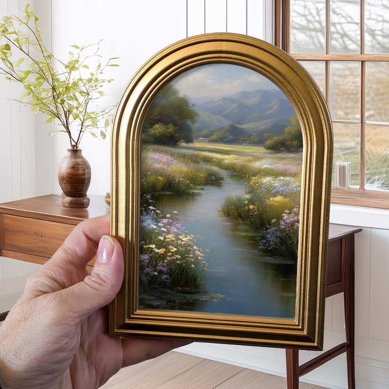 Pode incluir: Uma pintura de paisagem emoldurada com uma moldura dourada arqueada. A obra retrata uma serena cena de rio com flores silvestres coloridas, vegeta&ccedil;&atilde;o exuberante e montanhas distantes sob um c&eacute;u brilhante. A moldura adiciona um toque de eleg&acirc;ncia.