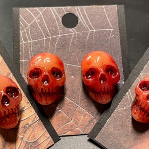 May include: Three red skull earrings with white eyes and teeth. The skulls are on a brown and black background.