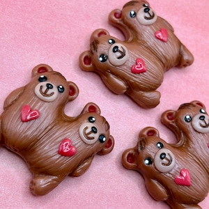 May include: Four brown bear-shaped decorative items with red heart accents. Each bear has a smiling face with black eyes and a light-colored snout. The bears are arranged on a pink background.