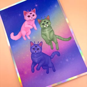 May include: A print featuring three alien cats in pink, green, and blue, each with three eyes and a crown-like antenna. The background is a gradient of purple, blue, and pink with stars. The print has a silver holographic border.