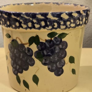 May include: A ceramic pot with a cream-colored base and a blue dotted rim. The pot is decorated with hand-painted clusters of purple grapes and green leaves, creating a rustic, farmhouse aesthetic.