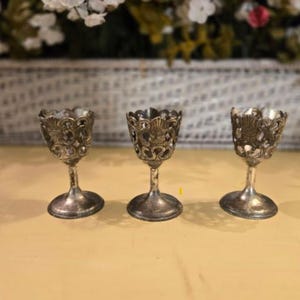 Puede incluir: Tres hueveras antiguas de color plateado con diseños calados ornamentados. Cada taza tiene un borde acampanado y decorativo y se asienta sobre una pequeña base redonda. Las hueveras están dispuestas sobre una superficie de color amarillo claro, con un arreglo floral borroso en el fondo.