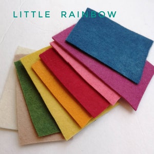 May include: A set of nine felt squares in various colours, including beige, green, yellow, orange, red, pink, purple, blue, and white. The squares are arranged in a rainbow pattern, with the white square at the top and the blue square at the bottom. The text "LITTLE RAINBOW" is written in green letters at the top of the image.