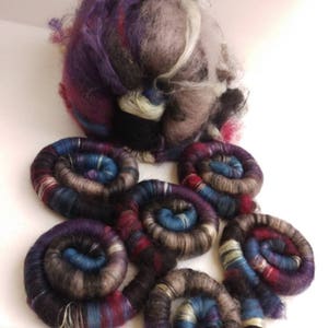 May include: A collection of hand-spun yarn in shades of purple, blue, grey, and white, arranged in a spiral pattern.