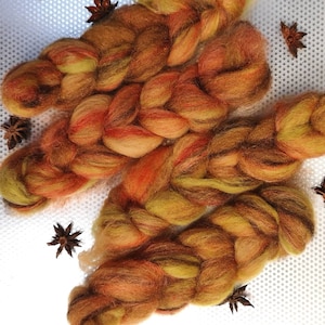 May include: Braided fiber art yarn in shades of orange, yellow, and brown. The yarn is arranged in three sections, with star anise scattered around. The yarn appears soft and fluffy, ideal for fiber art projects.