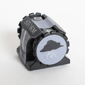 May include: A black weather die with a gray cloud and lightning bolt icon. The word "STORM" is printed on the side. The die has a rotating dial with numbers and weather symbols.