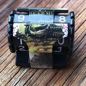May include: A black, mechanical counter with white numbers and the word "GERM" printed on top. The counter has a decorative sticker wrapped around it with a nature scene.
