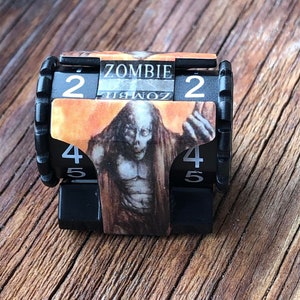 May include: Black plastic game counter with the word "ZOMBIE" and the numbers 2, 4, and 5. The counter has a picture of a zombie on it. The counter is on a wooden surface.