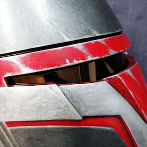 May include: A close-up of a grey and red helmet with a visor. The helmet has a weathered look with scratches and scuffs.