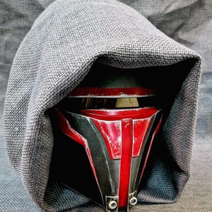 Könnte beinhalten: Nahaufnahme eines schwarz-roten Helms mit abgenutztem Aussehen, teilweise bedeckt von einer grauen Stoffhaube. Der Helm hat ein markantes Design mit einem zentralen roten Streifen und metallischen Akzenten, was auf ein Science-Fiction- oder Fantasy-Thema hindeutet.