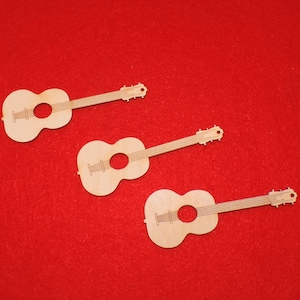 Może przedstawiać: Trzy małe, jasne wycięcia z drewna w kształcie gitary ułożone na czerwonym tle. Każda gitara ma okrągły otwór rezonansowy i długą szyjkę z kołkami do strojenia. Gitary są prawdopodobnie przeznaczone do rękodzieła lub dekoracji.