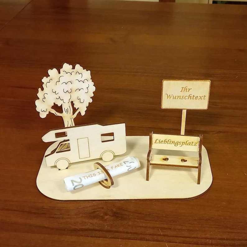 May include: A wooden model of a camper van, a tree, a bench, and a sign that says "Ihr Wunschtext" and "Lieblingsplatz". The camper van has a rolled up bill of money attached to it.