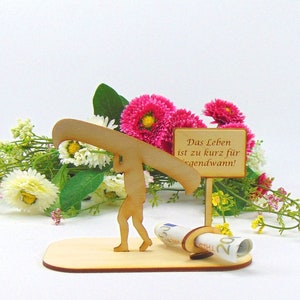 May include: A light-coloured wooden cutout of a person carrying a canoe on their shoulders, standing on a flat base. Next to them is a small sign with the text "Das Leben ist zu kurz f&uuml;r irgendwann!" and a circular holder containing a rolled banknote.