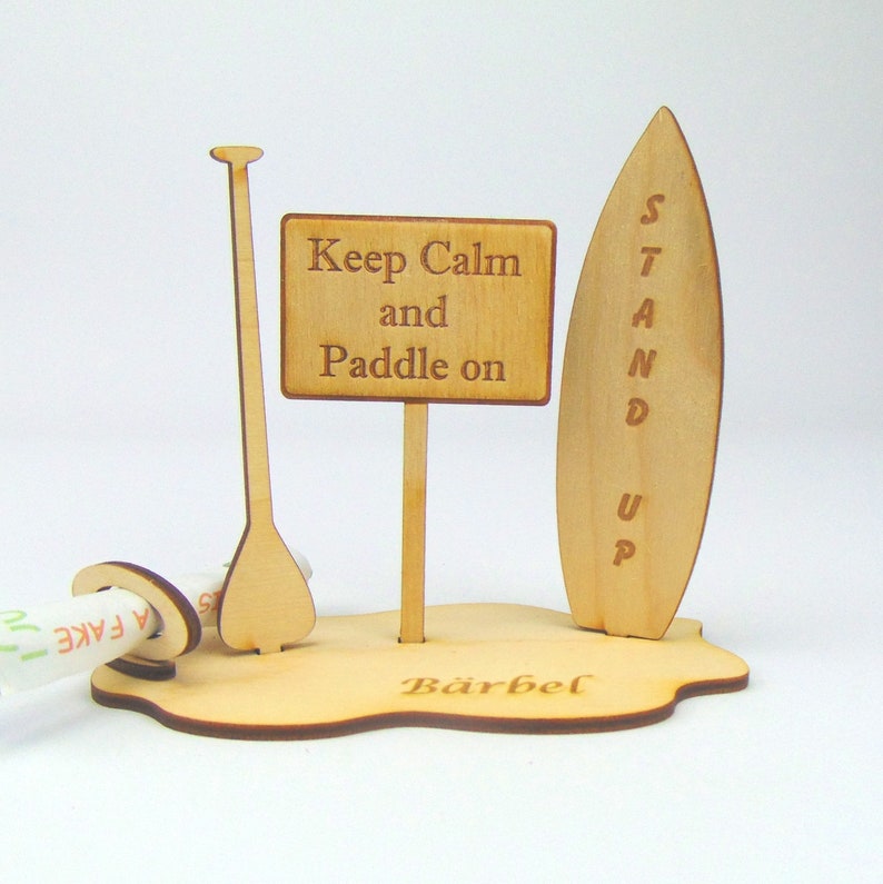 May include: Wooden stand up paddle board with a paddle and a sign that says "Keep Calm and Paddle On". The stand is personalized with the name "B&auml;rbel".