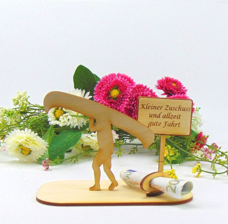 May include: A light-coloured wooden cutout featuring a figure carrying a canoe on their shoulders, standing on a flat base next to a sign that reads "Kleiner Zuschuss und allzeit gute Fahrt" and a rolled-up twenty euro banknote held in a small ring.