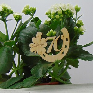 May include: A wooden horseshoe with the number 70 and the name Roberto engraved on it. The horseshoe is decorated with a four-leaf clover. The horseshoe is placed in a green plant.