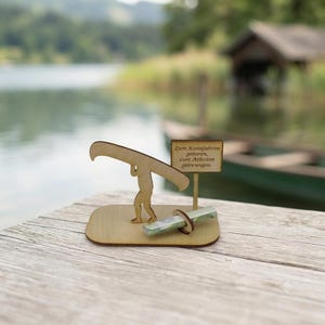 May include: A light-coloured wooden figurine of a person carrying a canoe on their shoulders, mounted on a flat base. Next to the figure is a small sign with the text 'Zum Kanufahren geboren, zum Arbeiten gezwungen' and a rolled-up paper scroll.