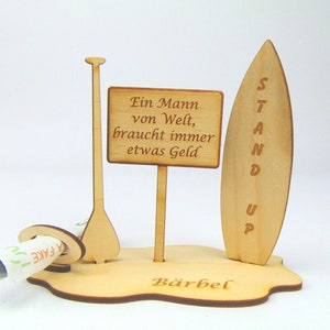 May include: Wooden stand up money box with a surfboard, paddle and a sign that reads "Ein Mann von Welt, braucht immer etwas Geld". The base of the stand is inscribed with the name "B&auml;rbel".