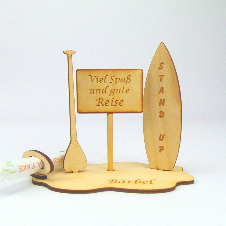 May include: A wooden cutout of a surfboard, paddle, and sign with the text "Viel Spa&szlig; und gute Reise" and the name "B&auml;rbel" at the bottom.