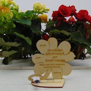 Op de afbeelding: Houten klavertje vier kaart met de tekst "Herzlichen Gl&uuml;ckwunsch zur Konfirmation" en "M&ouml;ge Gott dich begleiten und besch&uuml;tzen." De kaart is versierd met een hartvormige basis en een rol voor een gepersonaliseerd bericht.