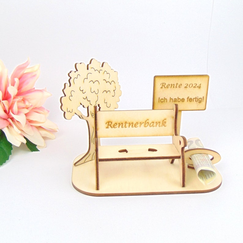 K&ouml;nnte beinhalten: Holzfigur einer Parkbank mit einem Baum und einem Schild mit der Aufschrift "Rente 2024 Ich habe fertig!" Die Bank ist mit "Rentnerbank" beschriftet.
