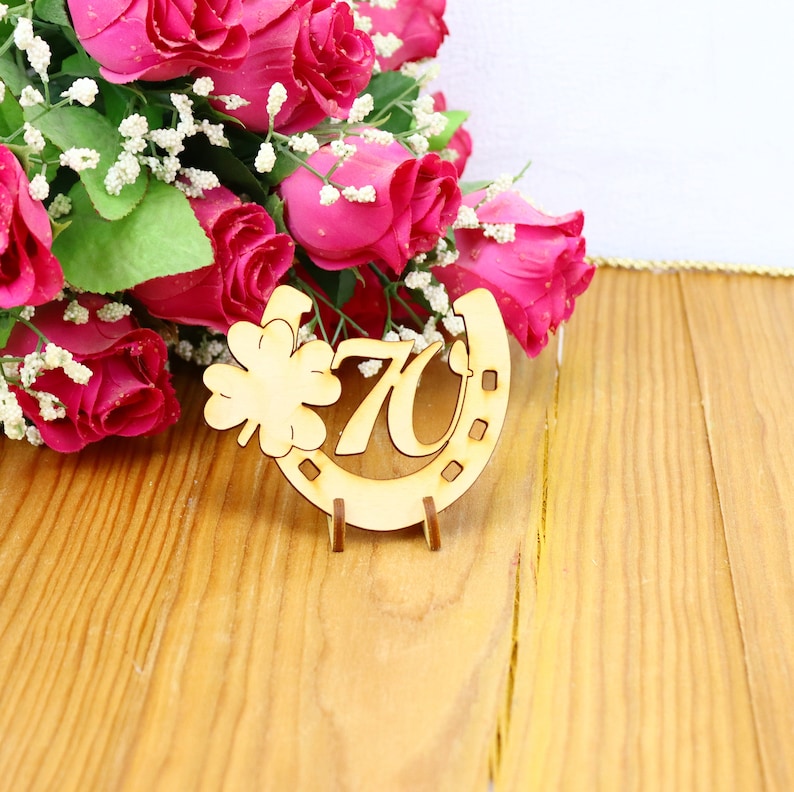 May include: A wooden horseshoe with the number 70 and a four-leaf clover design. The horseshoe is on a wooden surface with a bouquet of pink roses in the background.
