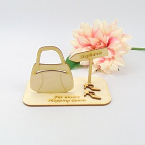 May include: Wooden decorative piece featuring a handbag, high heels, and a sign that reads "Stephanie." The base has the text "F&uuml;r unsere Shopping Queen." A pink and white flower is in the background.