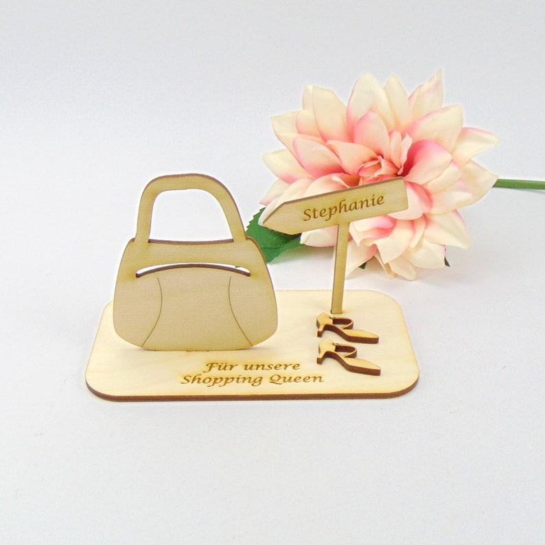 May include: Wooden decorative piece featuring a handbag, high heels, and a sign that reads "Stephanie." The base has the text "F&uuml;r unsere Shopping Queen." A pink and white flower is in the background.