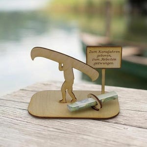 May include: A light-coloured wooden figurine of a person carrying a canoe on their shoulders, mounted on a flat base. Next to the figure is a small sign with the text "Zum Kanufahren geboren, zum Arbeiten gezwungen" and a circular holder for currency.