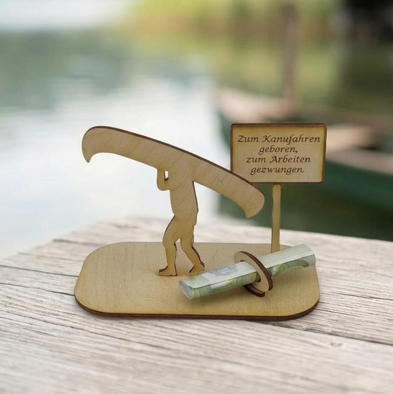May include: A light-coloured wooden figurine of a person carrying a canoe on their shoulders, mounted on a flat base. Next to the figure is a small sign with the text "Zum Kanufahren geboren, zum Arbeiten gezwungen" and a circular holder for currency.