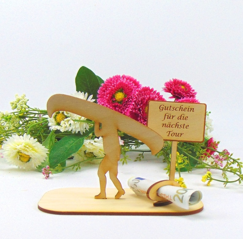 May include: A light-coloured wooden cutout featuring a figure carrying a canoe on their shoulders, standing on a flat base. Next to the figure is a rectangular sign with the text "Gutschein f&uuml;r die n&auml;chste Tour" and a circular holder containing a rolled banknote.