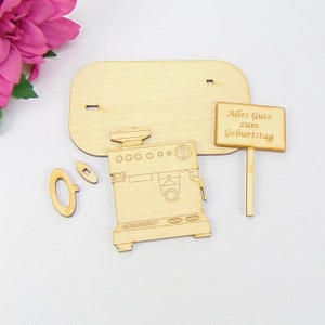 Pu&ograve; includere: Un kit per macchina da caff&egrave; espresso in legno con un cartello che recita "Alles Gute zum Geburtstag". Il kit include una base, parti della macchina e un piccolo cartello. I pezzi sono in legno chiaro.
