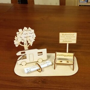 May include: A wooden model of a camper van, a tree, a bench, and a rolled-up bill of money. The bench has the words "Lieblingsplatz" on it. A sign above the camper van reads "Ruhestand Franz Mustermann 01.10.2019 Arbeiten".