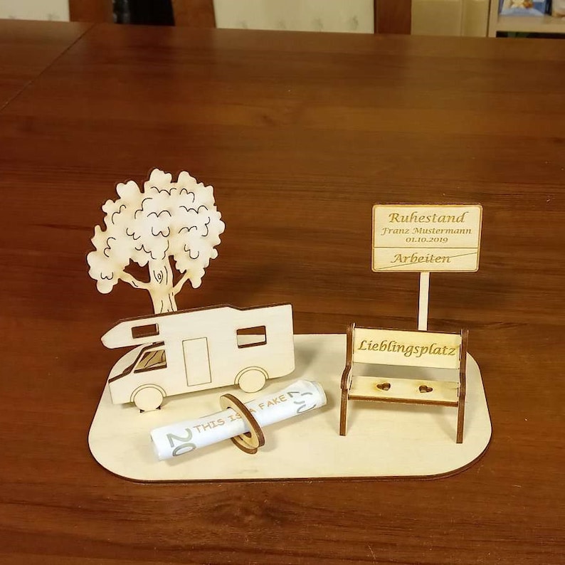May include: A wooden model of a camper van, a tree, a bench, and a rolled-up bill of money. The bench has the words "Lieblingsplatz" on it. A sign above the camper van reads "Ruhestand Franz Mustermann 01.10.2019 Arbeiten".