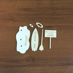 May include: A wooden craft kit for making a stand up paddle board. The kit includes a paddle, a board, a person, and a sign that says "Mir reicht's, ich geh paddeln".